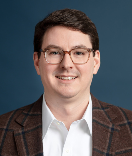 A man wearing glasses, a brown plaid blazer, and a white shirt smiles against a blue background.